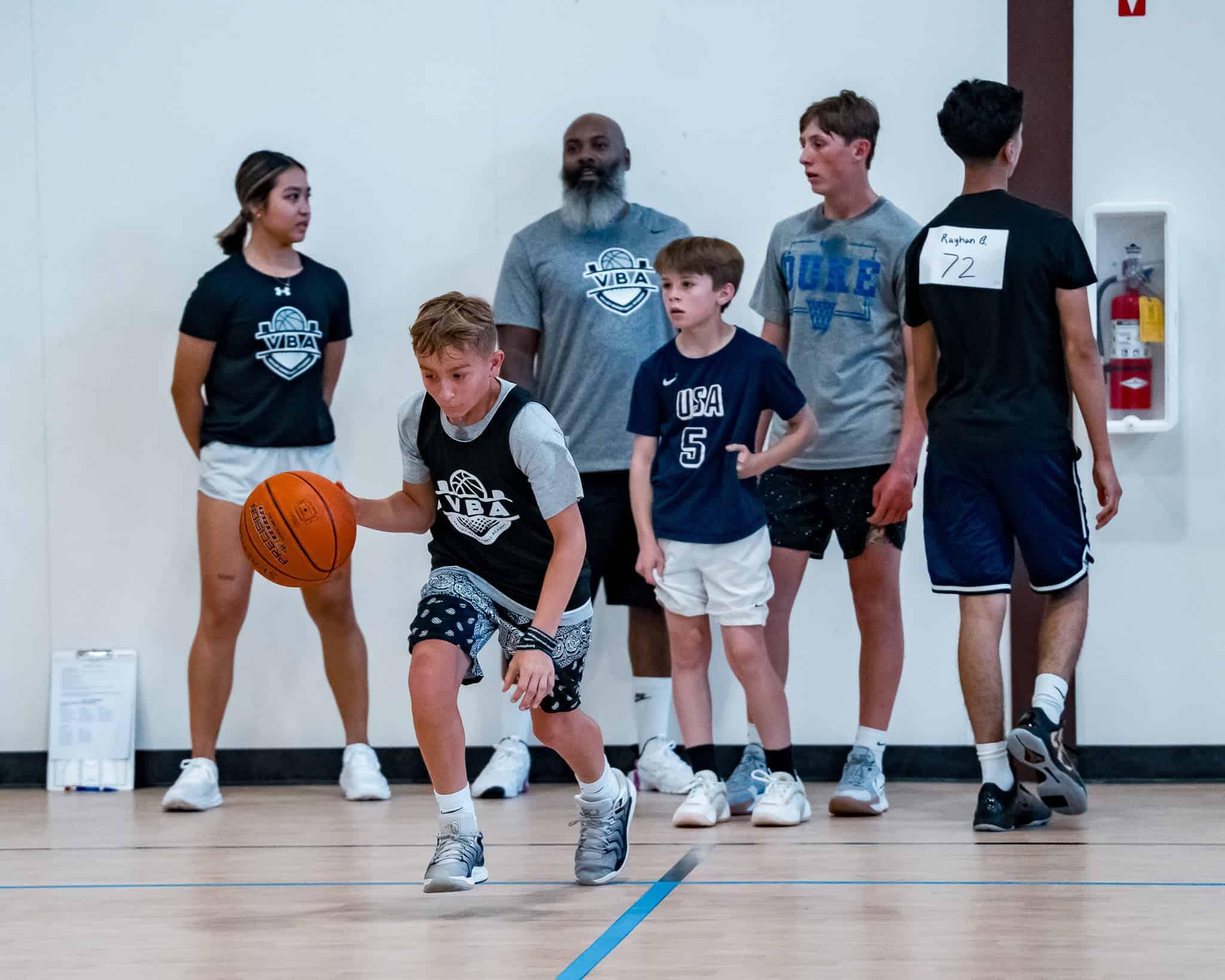 A youth basketball player driving to the basket during an AAU tryout while a coach evaluates from the sideline.
